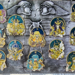 Könnte beinhalten: Zwölf goldfarbene Emaille-Pins mit Figuren aus der Anime-Serie Saint Seiya. Jeder Pin zeigt ein anderes Sternzeichen, wobei die Figur eine goldene Rüstung trägt. Die Pins sind in einem Kreis um ein zentrales Bild von zwei Frauenköpfen angeordnet.