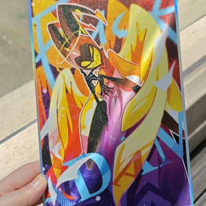 May include: A holographic sticker featuring a cartoon character with yellow wings, a red and black outfit, and a halo. The character is holding their hands together in front of their chest. The background is a colorful abstract design with the text "Frickin'" and "Damn" visible.