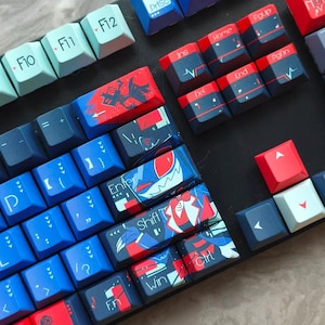 May include: A mechanical keyboard featuring a custom keycap set with a vibrant colour scheme of red, dark blue, and light blue. The keys display various graphic illustrations of characters, along with standard text labels like Enter, Shift, and Ctrl.