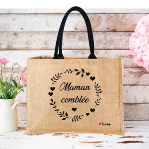 May include: A brown burlap tote bag with black handles. The bag has a black design with the text "Maman combl&eacute;e" and a wreath of leaves and hearts. The text "Clara" is written in red at the bottom of the design.