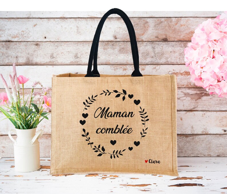 May include: A brown burlap tote bag with black handles. The bag has a black design with the text "Maman combl&eacute;e" and a wreath of leaves and hearts. The text "Clara" is written in red at the bottom of the design.
