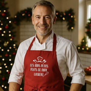 Peut inclure: Un homme porte une veste de chef blanche et un tablier rouge. Le tablier porte du texte blanc et un dessin de toque de chef. Le texte dit "LES BONS PETITS PLATS DE PAPA LAURENT". L'arrière-plan comprend un sapin de Noël et des décorations festives.