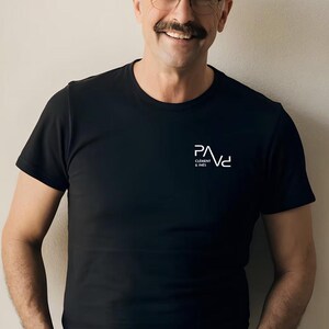 K&ouml;nnte beinhalten: Schwarzes T-Shirt mit Rundhalsausschnitt und dem wei&szlig;en Logo "PAVd CLEMENT & INES" auf der linken Brust. Das kurz&auml;rmelige Shirt ist aus einem weichen Material. Das Model tr&auml;gt eine Brille und hat einen Schnurrbart.