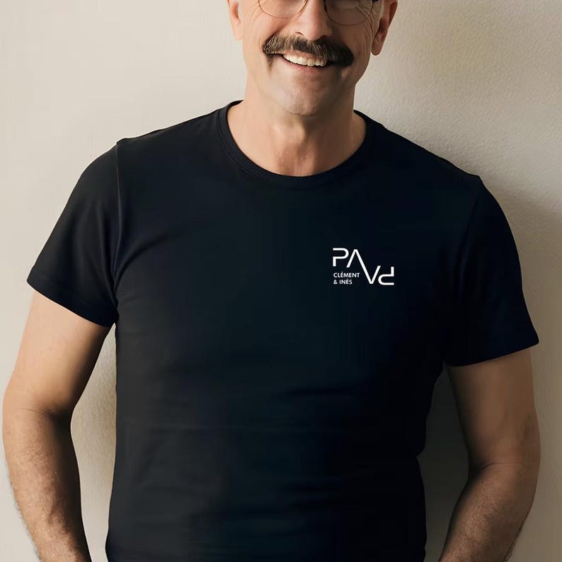 K&ouml;nnte beinhalten: Schwarzes T-Shirt mit Rundhalsausschnitt und dem wei&szlig;en Logo "PAVd CLEMENT & INES" auf der linken Brust. Das kurz&auml;rmelige Shirt ist aus einem weichen Material. Das Model tr&auml;gt eine Brille und hat einen Schnurrbart.