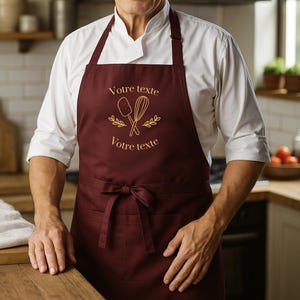 May include: A burgundy apron with the text "Votre texte" in gold, along with a whisk and spatula design. The apron has a front pocket and a bow at the waist. The person is wearing a white chef's coat.