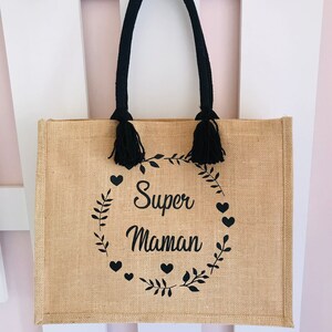 May include: A brown burlap tote bag with black handles and tassels. The bag has a black print of a wreath with leaves and hearts that says "Super Maman".