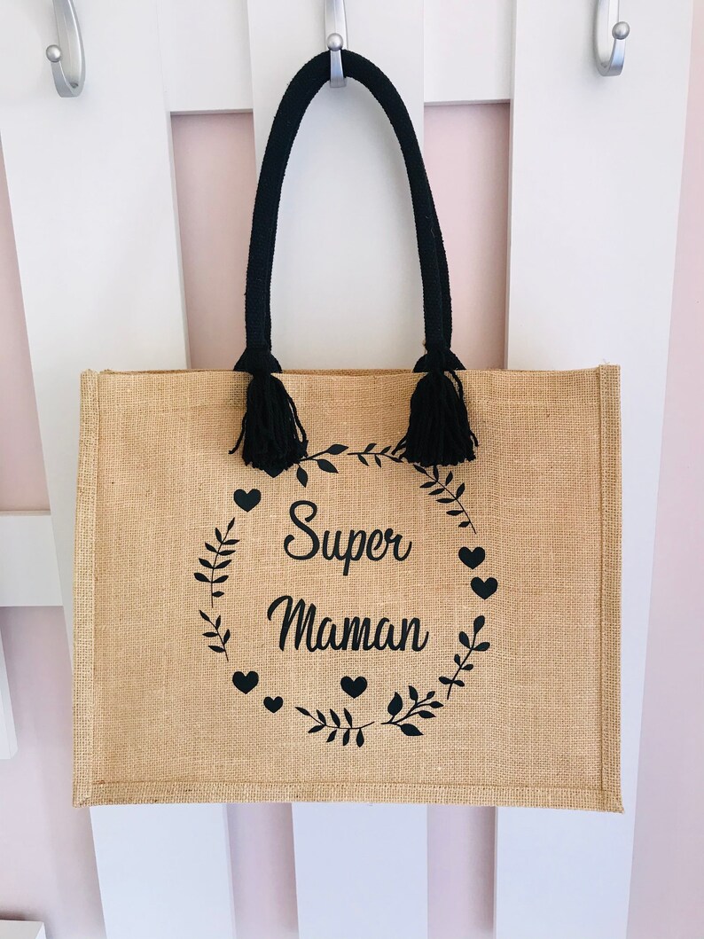 May include: A brown burlap tote bag with black handles and tassels. The bag has a black print of a wreath with leaves and hearts that says "Super Maman".