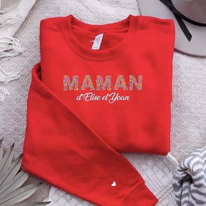 Peut inclure: Un sweat-shirt rouge avec le mot "MAMAN" en lettres fleuries et le texte "d'Elise et Yoan" en blanc. Un petit cœur blanc est sur la manche gauche. Le sweat-shirt est plié sur une surface à motifs.