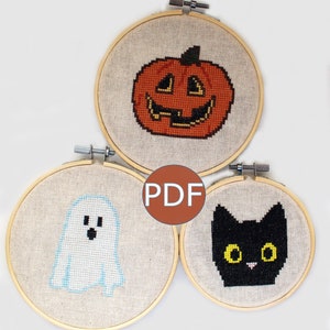 May include: Three embroidery hoops with Halloween-themed cross-stitch patterns: a smiling pumpkin, a ghost, and a black cat. The patterns are in a pixelated style. The hoops are made of wood and have a natural finish.