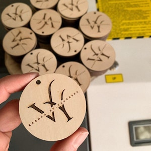 May include: A close-up of a wooden circle with the text "KASHPO BY TAVORSKA" and the letter "W" engraved on it. The circle is being held by a hand.