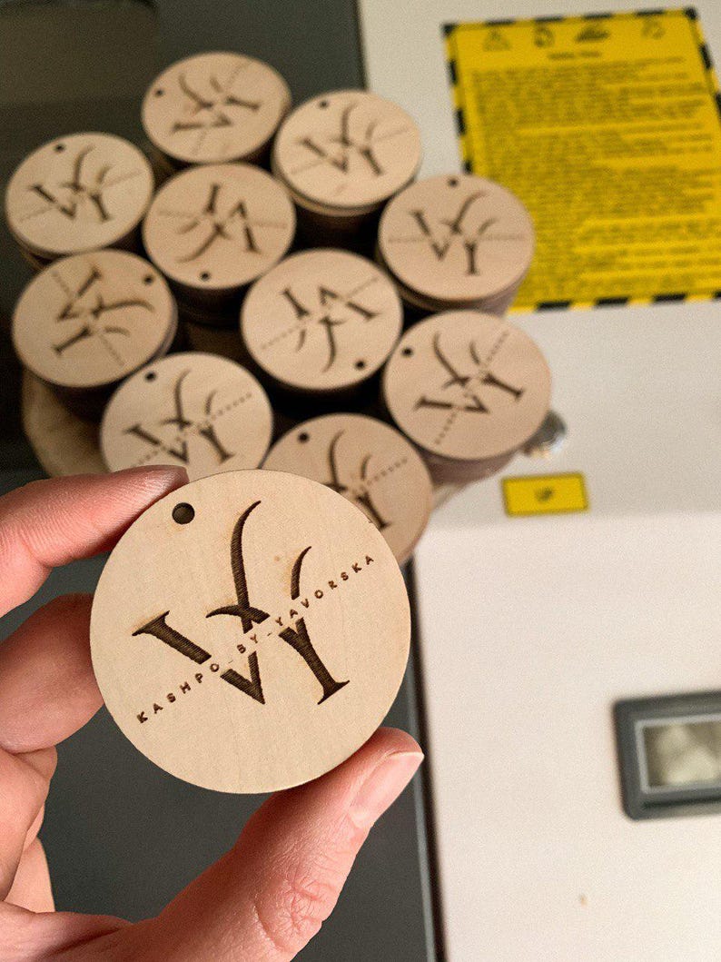 May include: A close-up of a wooden circle with the text "KASHPO BY TAVORSKA" and the letter "W" engraved on it. The circle is being held by a hand.