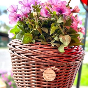 May include: A hanging wicker basket planter with pink petunias. The basket has a brown stain and a wooden tag with the text "@faina.kashpo.ua made in Ukraine 0632551260".