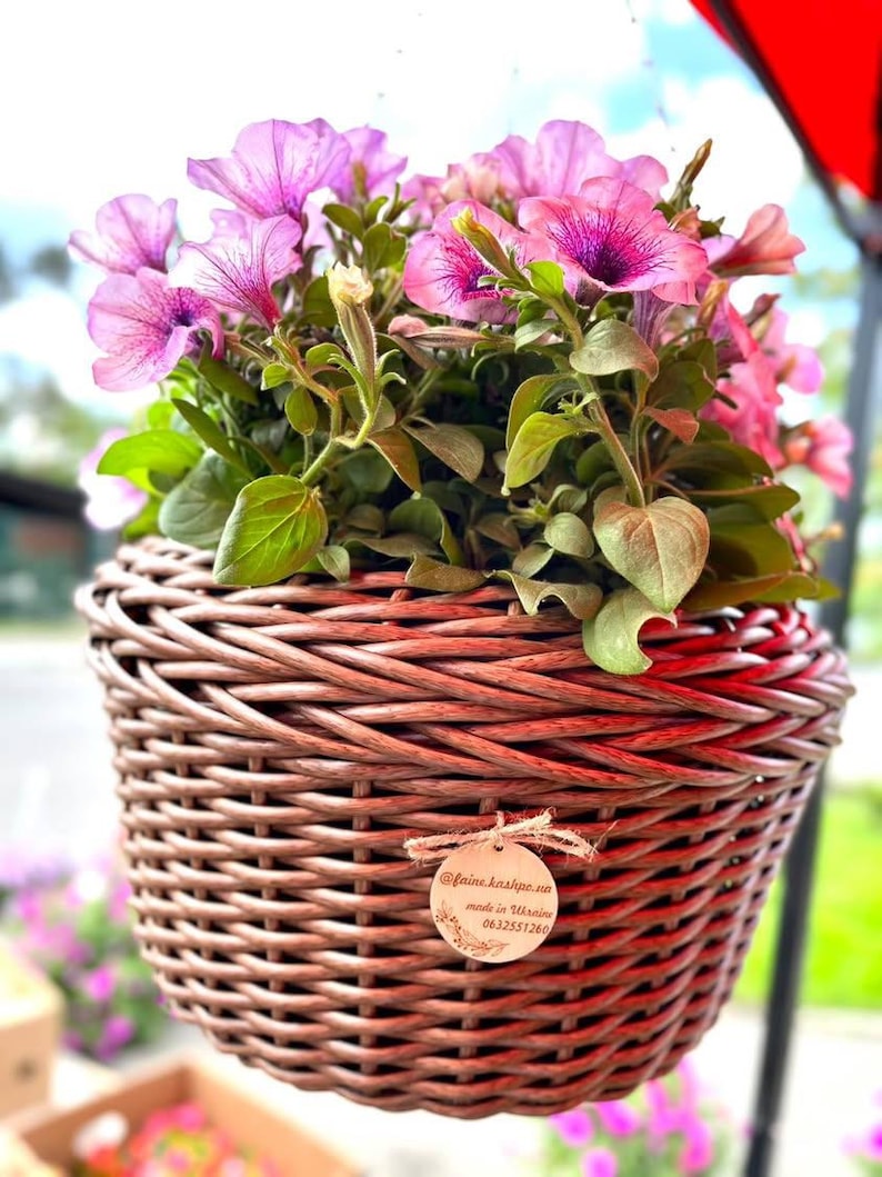 May include: A hanging wicker basket planter with pink petunias. The basket has a brown stain and a wooden tag with the text "@faina.kashpo.ua made in Ukraine 0632551260".