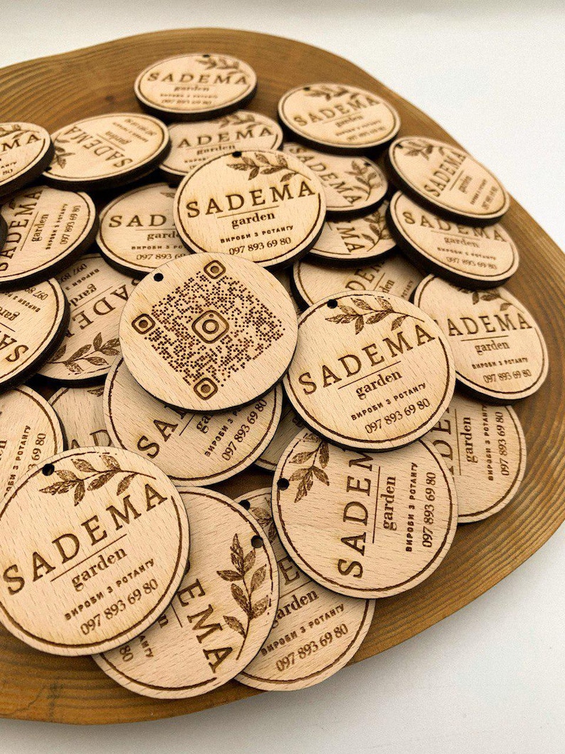May include: A close-up of a pile of round wooden tags with the text "SADEMA garden" and a phone number. The tags have a natural wood finish and are decorated with a small leaf design.