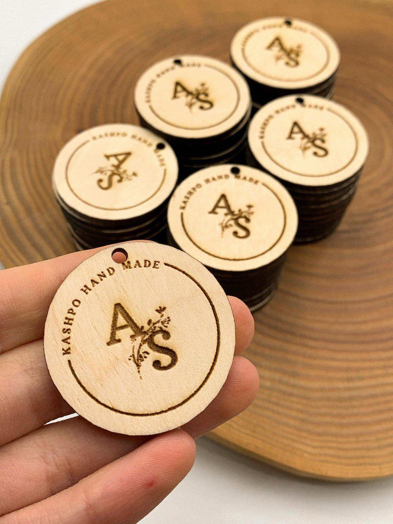 May include: A stack of round wooden tags with the letters "AS" and the words "KASHPO HAND MADE" engraved on them.