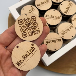 Puede incluir: Un conjunto de etiquetas redondas de madera clara, cada una con un agujero. Una etiqueta muestra un código QR, otras presentan el texto "Mr. Ralph Beautiful Lighting". Las etiquetas son de color marrón claro y parecen cortadas con láser.