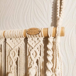 May include: A macrame wall hanging with a wooden dowel at the top. The hanging features a variety of knot patterns and has tassels at the bottom. The hanging is made with natural colored cotton cord. A wooden tag with the text "Instagram: @bahroma_store, Hand made with love" is attached to the dowel.