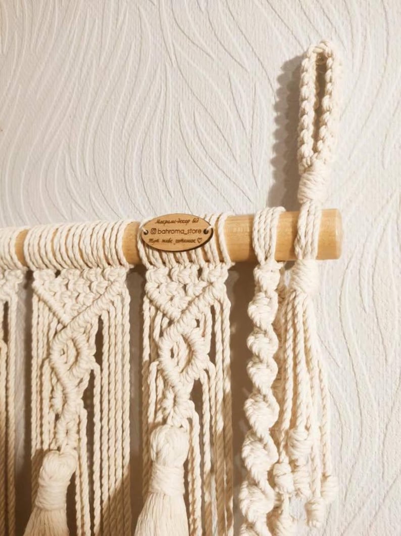 May include: A macrame wall hanging with a wooden dowel at the top. The hanging features a variety of knot patterns and has tassels at the bottom. The hanging is made with natural colored cotton cord. A wooden tag with the text "Instagram: @bahroma_store, Hand made with love" is attached to the dowel.