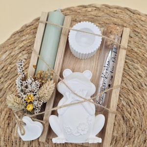 Könnte beinhalten: Ein dekoratives Holztablett mit einer hellgrünen Kerze, einer weißen Froschfigur, einem kleinen weißen Herz und einem Glasfläschchen mit Text. Das Tablett enthält auch Trockenblumen und einen weißen Kerzenhalter. Die Artikel sind mit Bindfaden zusammengebunden.