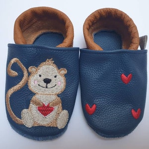 Könnte beinhalten: Ein Paar blaue Lederschuhe für Babys mit braunem Lederbesatz. Ein Schuh hat einen gestickten Affen, der ein rotes Herz hält, der andere hat drei gestickte rote Herzen.