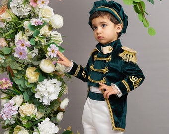 May include: A child's dark green velvet military-style suit with gold detailing, a matching hat, and white trousers. The child stands near a floral archway with white, peach, and pink flowers. Cream-coloured shoes complete the outfit.
