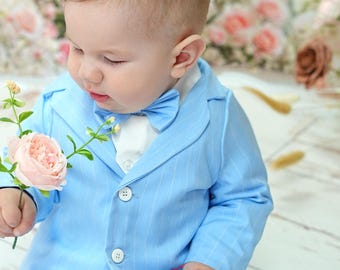 May include: A baby in a light blue pinstripe suit, bow tie, and white shirt, holding a pink flower. A matching hat sits nearby. The suit has two white buttons. The background features a floral arrangement.