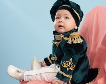 May include: A baby wearing a green velvet suit with gold trim and a matching hat sits on a pink chair. The suit has a gold embroidered design on the sleeves and a gold tassel on the collar.