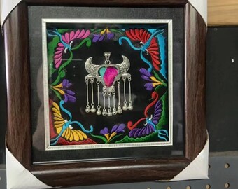 May include: A silver-toned metal pendant featuring a central pink and teal inlay, shaped like a stylized bull with dangling chains and small spherical charms, displayed inside a square dark wood frame with colorful floral embroidery.