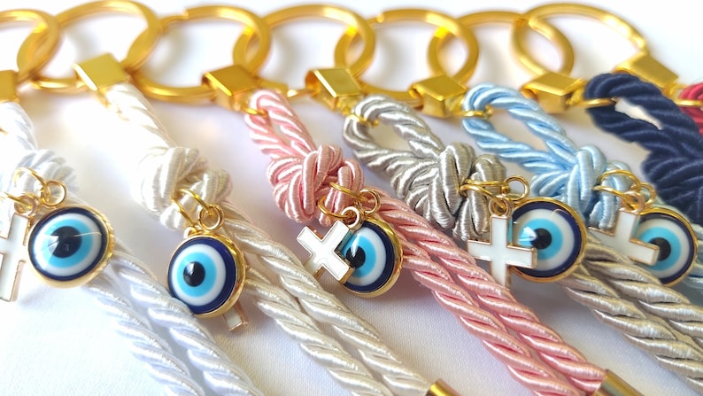May include: A set of six keychains with a gold ring and a braided cord. Each keychain has a charm with a blue evil eye and a white cross. The cords are in different colors: white, pink, gray, light blue, navy blue, and red.