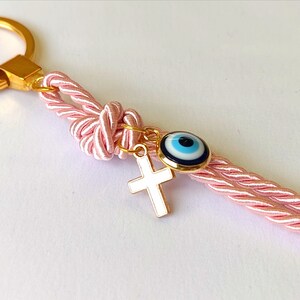 May include: Pink and gold keychain with a blue evil eye charm and a white cross charm. The keychain is made of a braided rope with gold accents.
