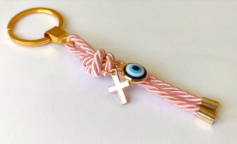 May include: Pink and gold keychain with a blue evil eye charm and a white cross charm. The keychain is made of a braided rope with gold accents.