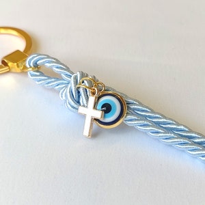 May include: A light blue braided keychain with a gold key ring, a gold cross charm, and a blue evil eye charm.