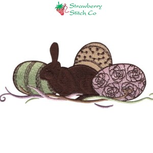 Puede incluir: Diseño bordado de un conejo marrón sentado en un nido de tres huevos de Pascua. Los huevos están decorados con rayas verdes, puntos marrones y rosas rosas.