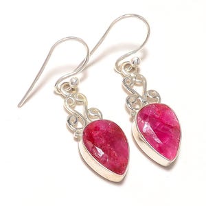 Może przedstawiać: Para srebrnych kolczyków wiszących z czerwonymi rubinowymi kamieniami szlachetnymi. Kolczyki mają delikatny, wirujący wzór.