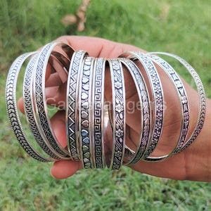 Op de afbeelding: Een set van negen zilveren armbanden met verschillende gegraveerde ontwerpen. De armbanden zijn op elkaar gestapeld en hebben een glanzende afwerking.