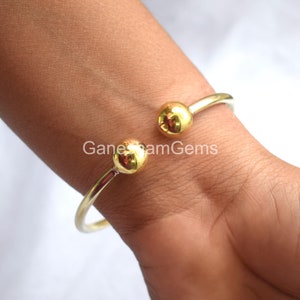 May include: A gold bangle bracelet with two round, gold balls on the ends. The bracelet is open and is being worn on a person's wrist.