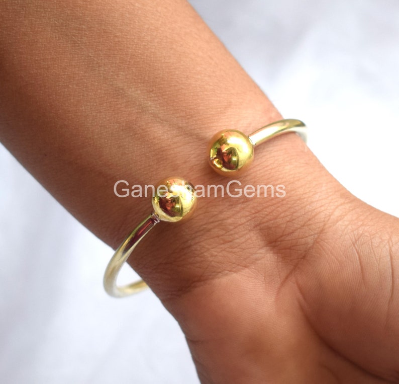 May include: A gold bangle bracelet with two round, gold balls on the ends. The bracelet is open and is being worn on a person's wrist.