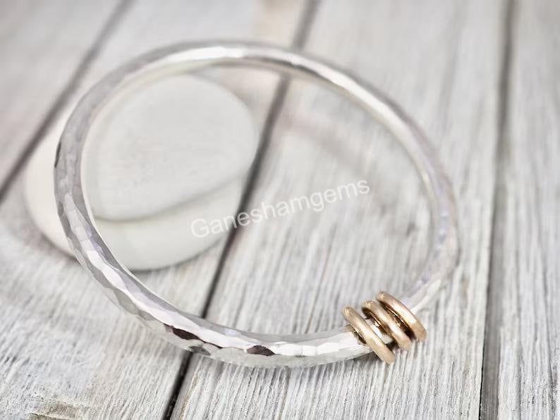 Peut inclure: Un bracelet en argent martel&eacute; avec une pile de quatre anneaux en or attach&eacute;s. Le bracelet est sur une surface en bois blanc.