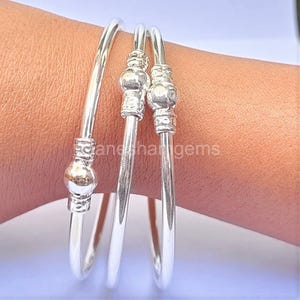 Peut inclure: Trois bracelets en argent sont pr&eacute;sent&eacute;s sur un poignet. Deux bracelets ont des accents d&eacute;coratifs perl&eacute;s, tandis que le troisi&egrave;me a un seul accent en forme de boule. Les bijoux sont brillants et r&eacute;fl&eacute;chissants, avec un design simple et &eacute;l&eacute;gant.
