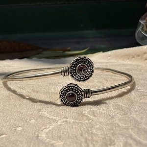 May include: A silver-toned cuff bracelet with two circular, textured accents. Each accent features a dark red gemstone at its center. The bracelet has an open design, allowing for an adjustable fit. The bracelet is displayed on a white surface.