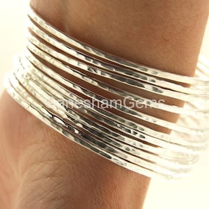 Puede incluir: Un conjunto de 12 pulseras de plata martilladas. Las pulseras están apiladas en la muñeca de una persona.