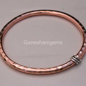 May include: A hammered copper bangle with a silver accent. The bangle is open and has a hammered texture. The silver accent is a small ring that is attached to the bangle.