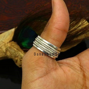 Könnte beinhalten: Ein silberner Spinner-Ring mit geflochtenem Seildesign und mehreren Bändern. Der Ring wird an einem Finger getragen und zeigt seine komplizierten Details und seine Handwerkskunst. Der Ring hat den Text "Ganesham Gems".