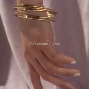 May include: A hand wearing multiple gold bangles. The bangles are smooth and shiny, reflecting light. The image includes the text "Ganesham Gems". The background is a soft, muted purple.