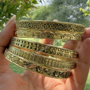 Könnte beinhalten: Eine Sammlung goldfarbener Armreifen mit aufwendigen floralen und geometrischen Mustern. Die Armreifen sind gestapelt und zeigen ihre detaillierten Designs und Texturen. Das Bild ist gut beleuchtet und hebt den metallischen Glanz des Schmucks hervor.