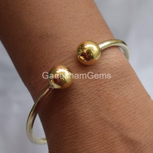 May include: A silver bangle bracelet with two gold ball accents. The bracelet is open and has a simple design.