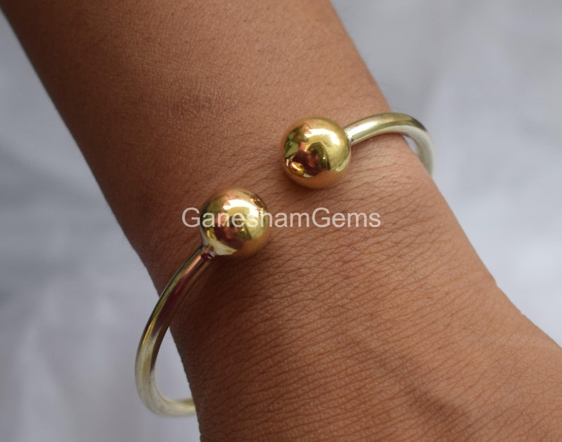 May include: A silver bangle bracelet with two gold ball accents. The bracelet is open and has a simple design.