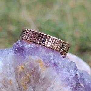 May include: A textured rose gold ring with a carved design. The ring is displayed on a purple crystal. The ring has a band with vertical lines. The word "Ganesh" is visible on the crystal.