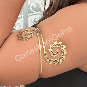 May include: A gold-colored arm cuff with spiral and geometric designs. The cuff features a curved bar that wraps around the upper arm, with decorative spiral elements at each end. The text "GaneshamGems" is visible on the image.