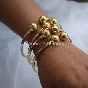 May include: Gold bangle bracelets with round ball accents. The bracelets are stacked on a person's wrist.